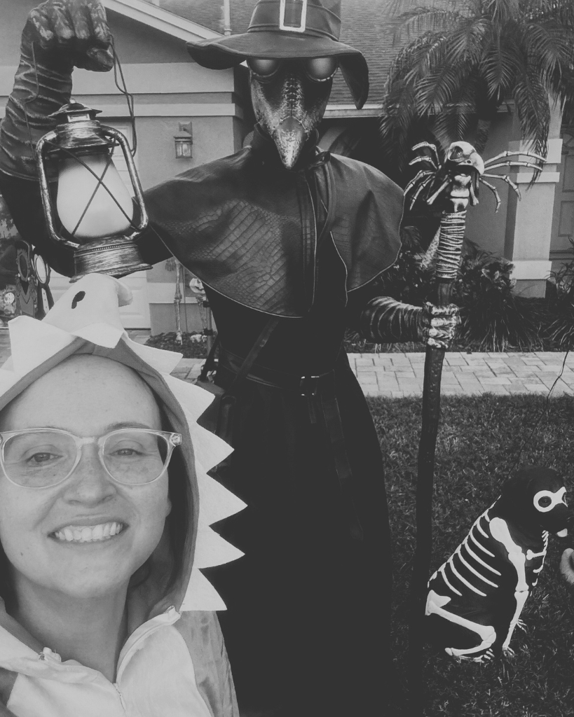 A playful Halloween-themed photo featuring a woman in a dinosaur costume smiling alongside a figure dressed as a plague doctor holding a lantern, with a skeleton dog decoration in the background.