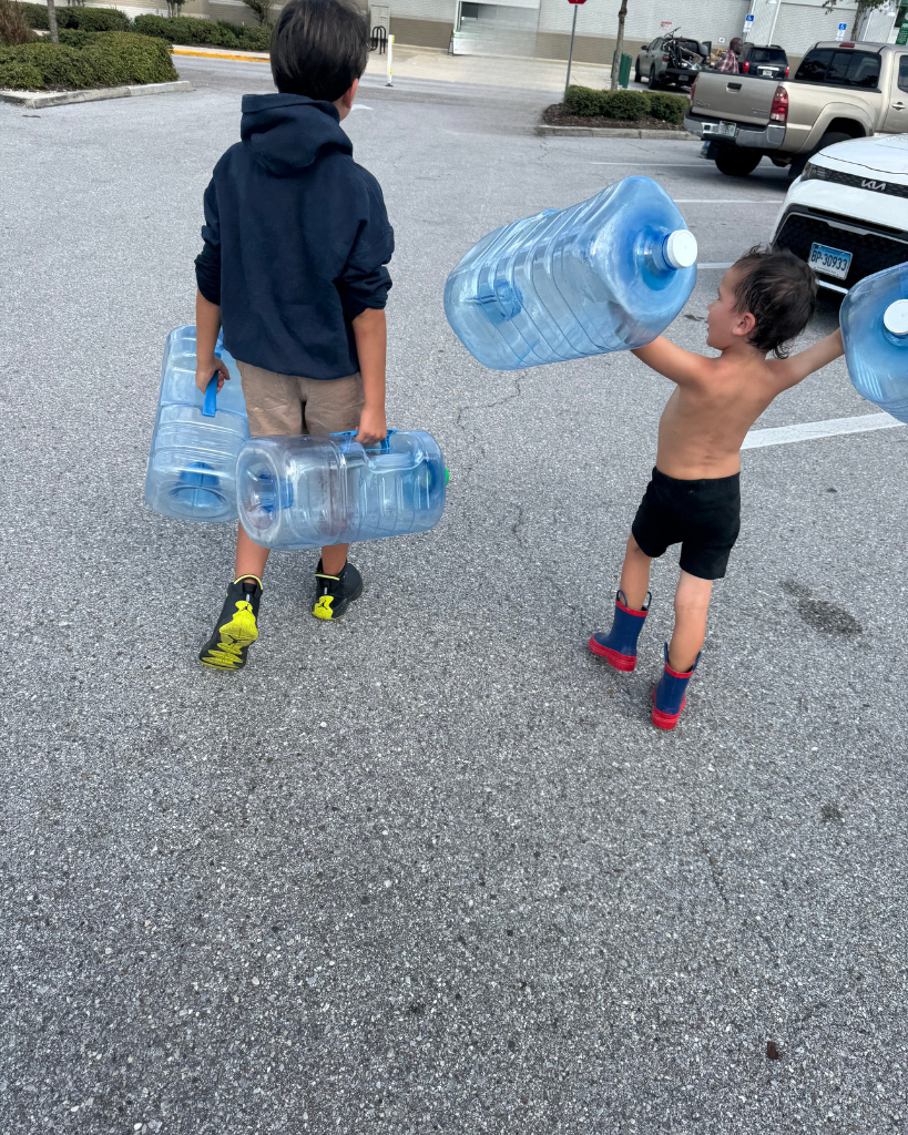 Two children carrying large water jugs in a parking lot, representing hard work, teamwork, and determination.