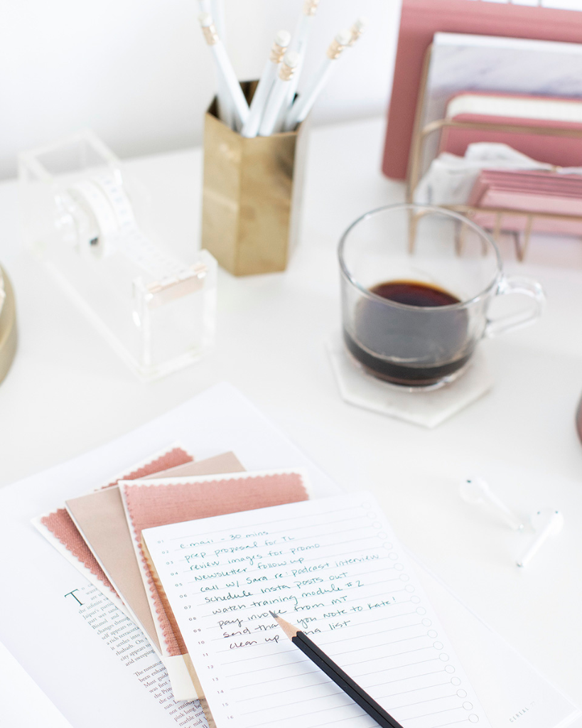 Organized workspace with a coffee cup, a written to-do list, notepads, pencils in a gold holder, and office supplies on a white desk.