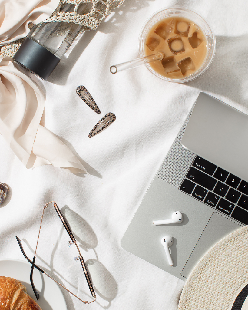 Flatlay of a productive morning setup featuring a laptop, iced coffee with a glass straw, AirPods, sunglasses, a croissant, and accessories on a white linen background.