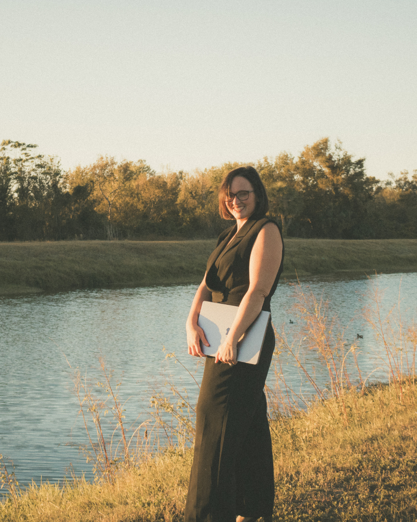 Jess Lazer standing near a peaceful lakeside, holding a laptop, symbolizing clarity and streamlined business growth.