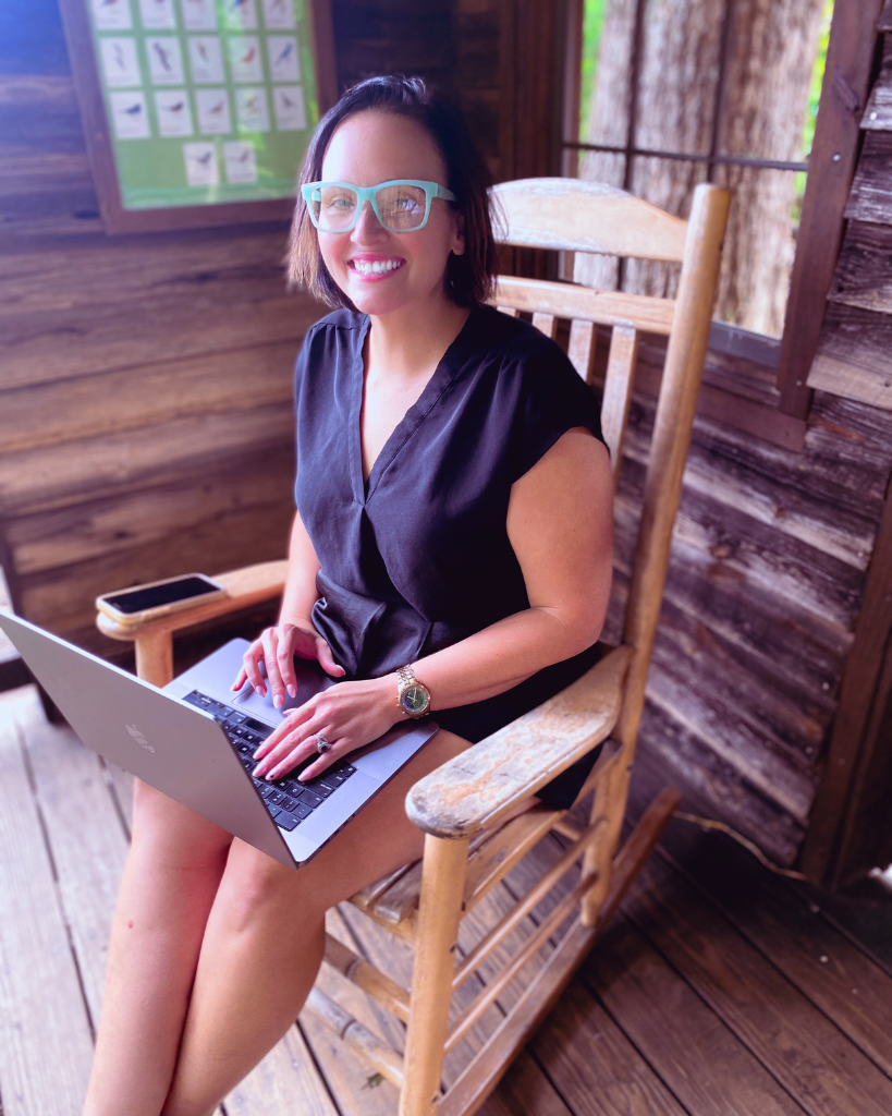 A smiling Jess Lazer sitting on a wooden rocking chair in a rustic cabin, working on her laptop, embodying focus and clarity.